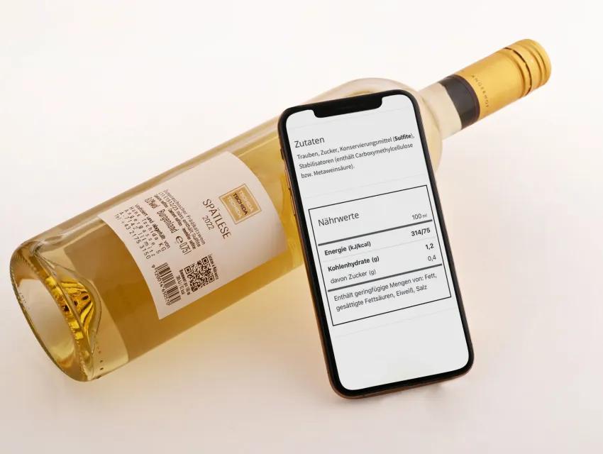 Foto einer Weinflasche mit E-Label und einem Smartphone, auf dessen Bildschirm Zutaten und Nährwerte des Weins zu sehen sind