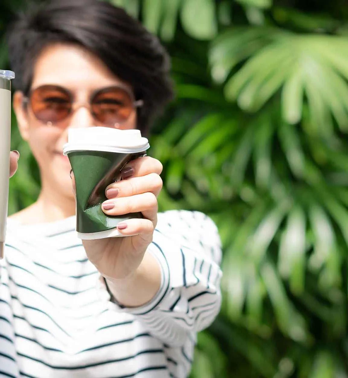 Frau hält einen wiederverwendbaren Coffee-to-go-Becher und einen zerdrückten Einweg-Becher in die Kamera